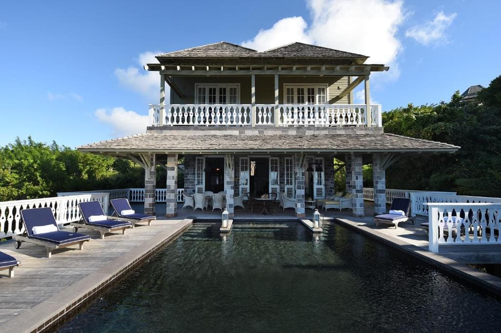 Two-story villa with a wraparound balcony, wooden roof, and white railings, featuring a rectangular pool surrounded by blue lounge chairs, set in a lush, green landscape under a blue sky with clouds.