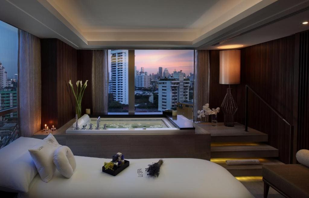 A luxurious spa room with a massage bed, a large bathtub by a window overlooking a city skyline at sunset, soft lighting, and elegant decor with flowers and candles.
