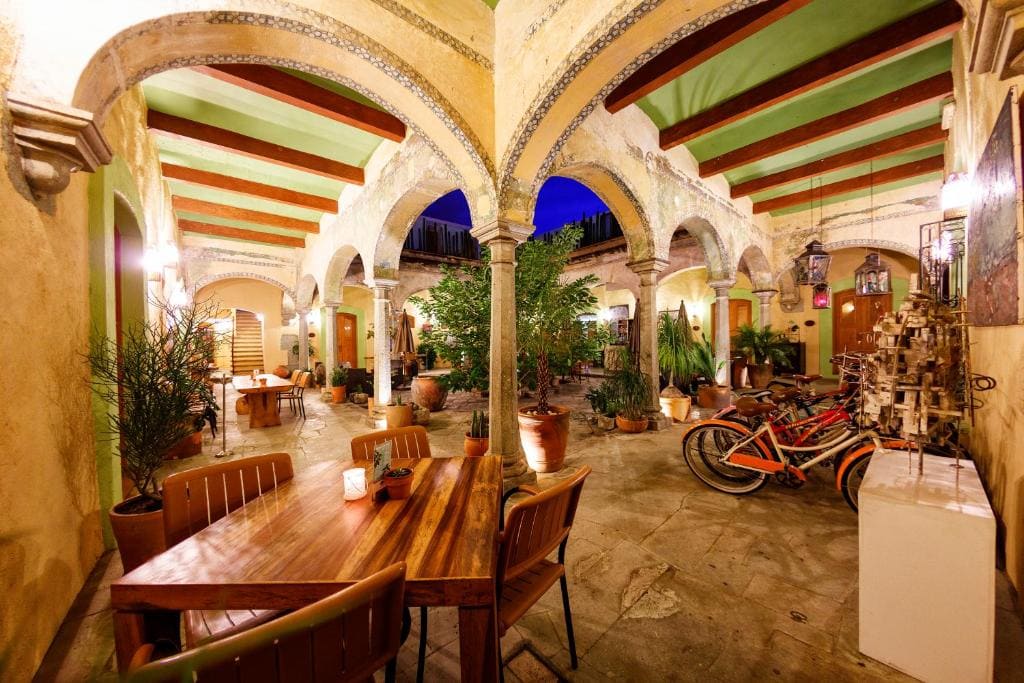A warmly lit courtyard with arched columns, potted plants, wooden tables and chairs, and several orange bikes parked near the wall. The ceiling has exposed beams, and the ambiance is cozy and inviting.
