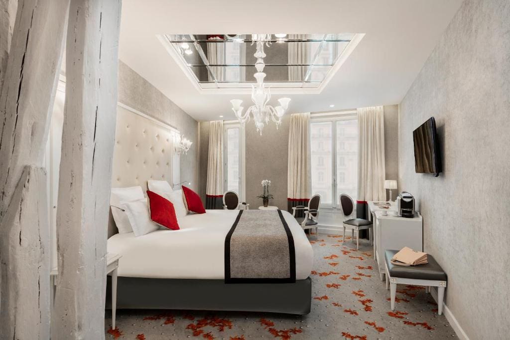 Elegant hotel room with a large bed, white and red pillows, gray walls, mirrored ceiling, chandelier, TV on the wall, two windows with curtains, desk, chairs, and floral carpet.