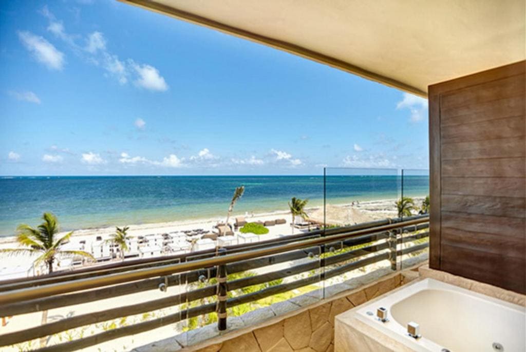 A beachfront balcony with a jacuzzi overlooking a sandy beach, palm trees, lounge chairs, and a clear blue ocean under a sunny sky.