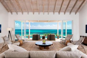 A modern living room with neutral furnishings opens to a patio, overlooking turquoise ocean and blue sky through large glass doors. Wicker chairs and greenery accent the airy, beachside setting.