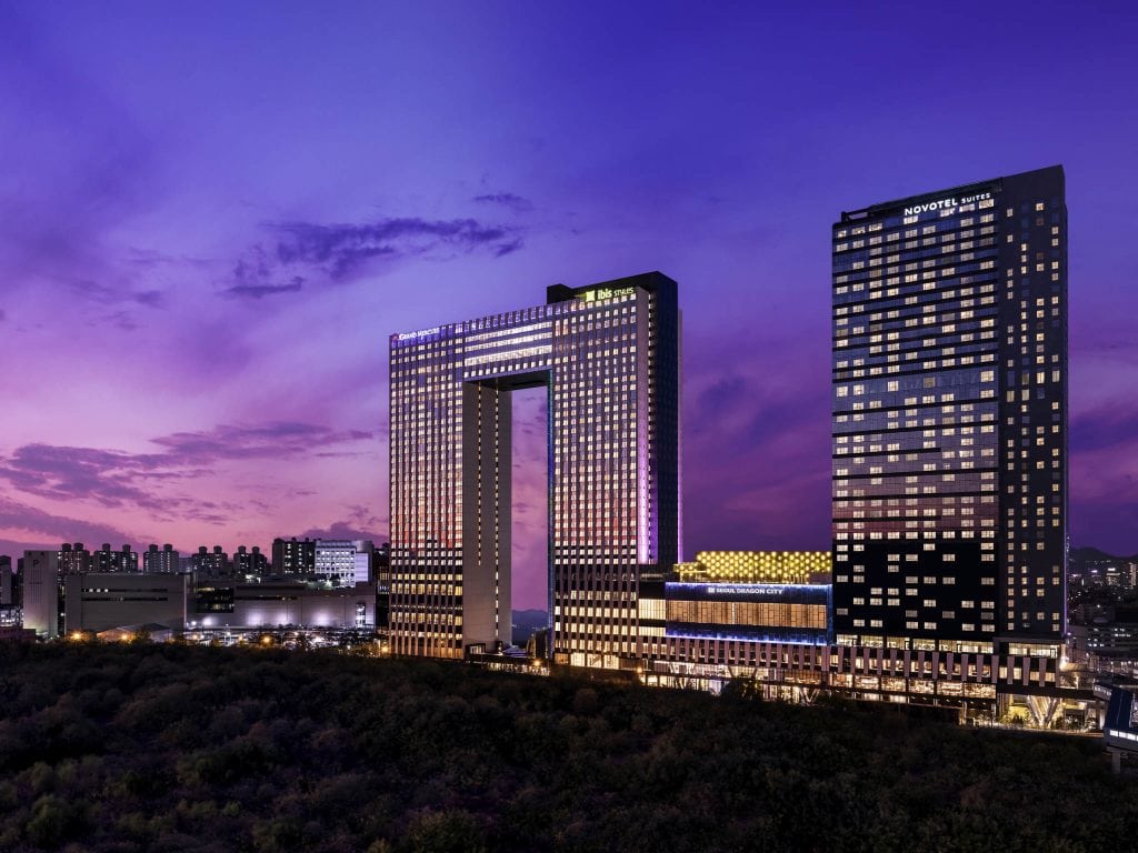 A modern cityscape at dusk, featuring two illuminated high-rise buildings, one with a large rectangular opening in the center, against a purple and pink sky. The foreground shows trees and the distant skyline.
