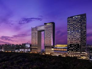 A modern cityscape at dusk, featuring two illuminated high-rise buildings, one with a large rectangular opening in the center, against a purple and pink sky. The foreground shows trees and the distant skyline.