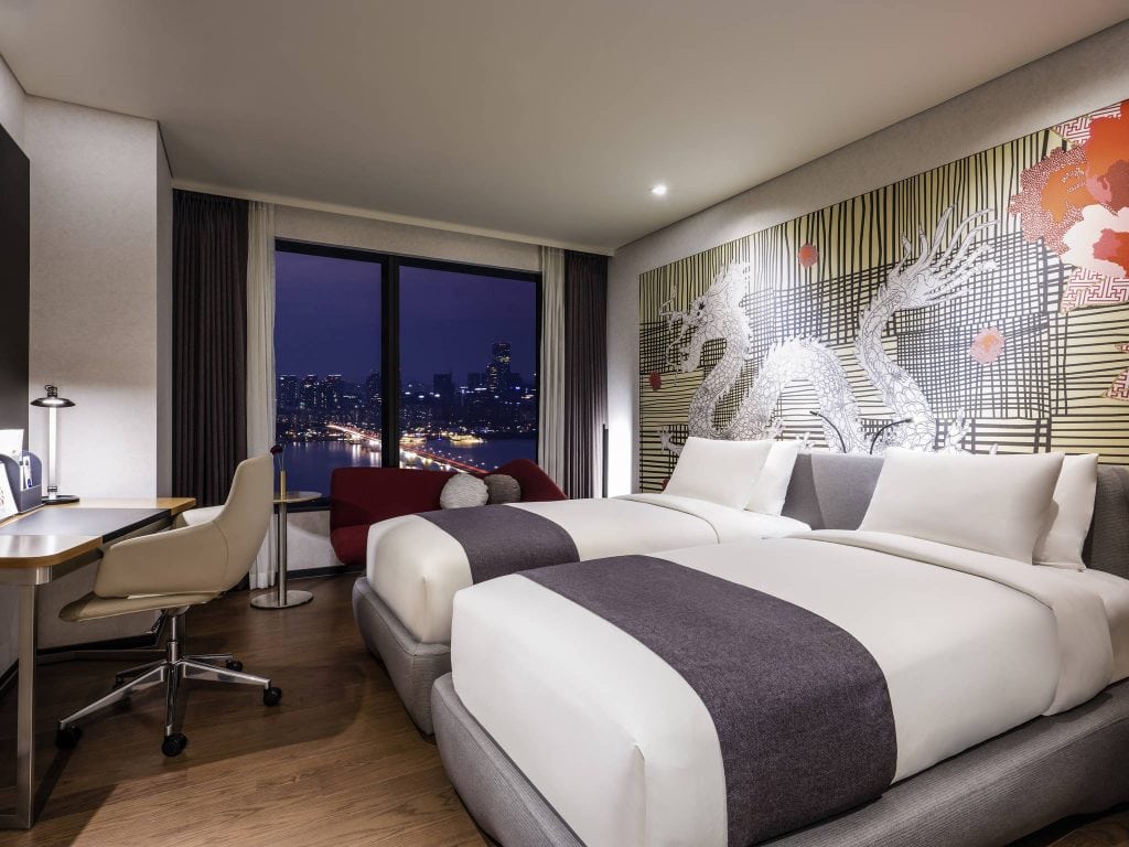 Modern hotel room with two double beds, gray and white bedding, a desk with a chair, a cityscape view through a large window at night, and a decorative dragon mural on the wall above the beds.