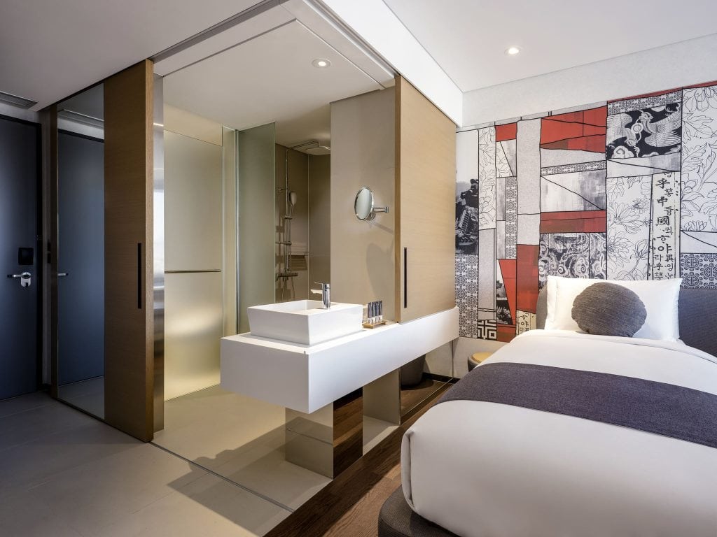 A modern hotel room features a double bed with white and gray bedding, a unique open bathroom with a floating sink, glass shower, and a colorful mural with artistic patterns on the wall beside the bed.