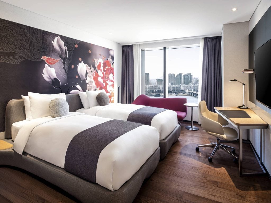 Modern hotel room with two twin beds, a floral accent wall, a desk with a chair, a maroon chaise lounge, wood floors, and a large window overlooking a city skyline and waterfront.