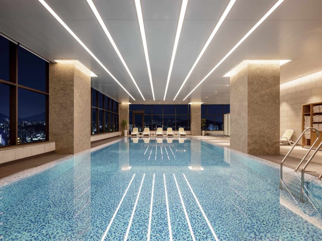 An indoor swimming pool with clear water, mosaic tiles, lounge chairs lined up at the far end, large windows showing a city night view, and modern ceiling lights illuminating the space.