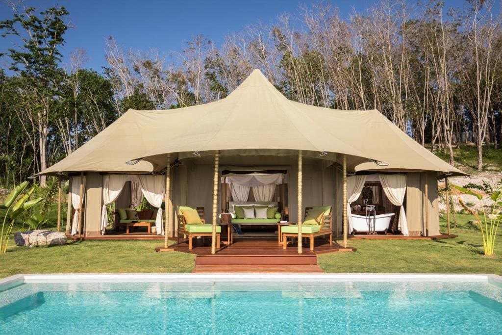 A luxurious glamping tent with a canopy roof, outdoor seating, and curtains stands on a wooden deck beside a clear swimming pool. Green grass and tall trees are in the background under a bright sky.