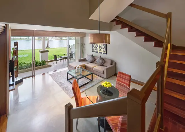 Modern living room with a gray sofa, glass coffee table, and dining area with orange chairs, next to large windows overlooking a lake and a patio with outdoor seating; wooden staircase on the right.