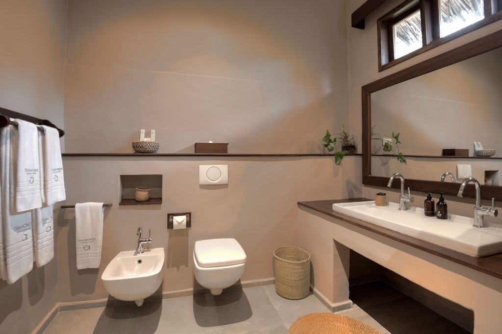 Modern bathroom with beige walls, floating toilet and bidet, large mirror above a double sink, white towels on a rack, woven basket, and potted plant on the counter for decoration.