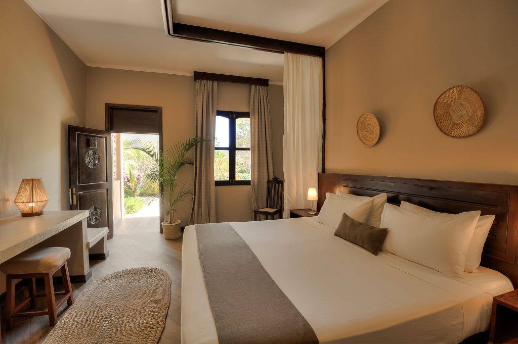 A cozy, sunlit bedroom with a large bed, neutral bedding, wooden furniture, woven wall decor, and an open door revealing greenery outside. Natural light fills the room, enhancing its warm and inviting atmosphere.