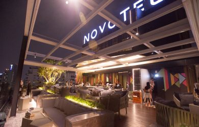 A modern rooftop bar at night with people sitting on sofas, greenery lining the area, and large illuminated NOVOTEL sign overhead; city lights visible in the background.