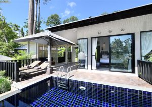 Modern villa with a private blue-tiled pool, wooden deck with lounge chairs, large umbrella, and sliding glass doors opening to a bright bedroom, surrounded by greenery under a clear blue sky.