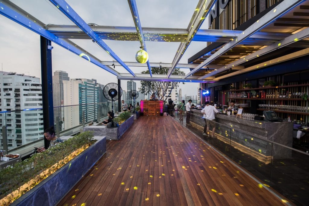 A rooftop bar with wooden flooring, lined planters, and a modern open roof, overlooks a city skyline. People are seated and standing near the bar, with lights and a disco ball creating a festive atmosphere.