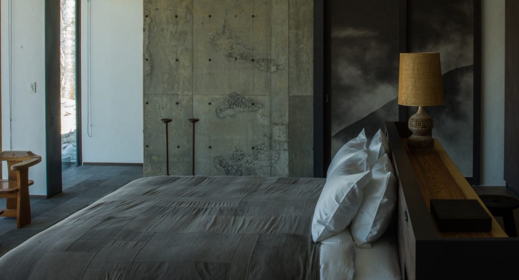 A modern, minimalist bedroom with a gray bed, concrete accent wall, two tall candle holders, a wooden chair, and a bedside lamp. Sunlight comes through a door on the left, illuminating part of the room.