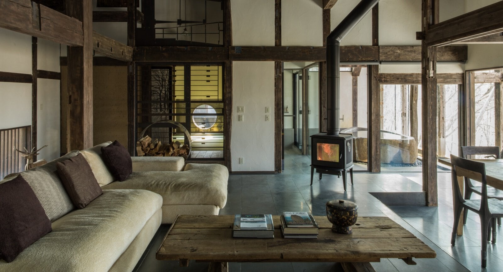 Spacious rustic living room with exposed wooden beams, beige sofa with dark pillows, wood-burning stove, wooden coffee table, and large windows letting in natural light. Firewood is stacked beside the stove.