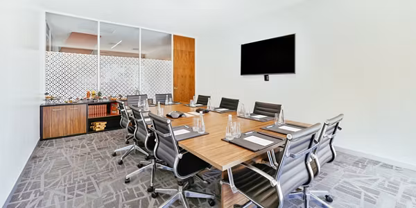 A modern conference room with a long wooden table, eight black chairs, placemats with glasses, a wall-mounted TV, and a sideboard with refreshments. Glass wall separates the room from another area.
