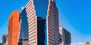 Tall modern office buildings with reflective glass windows and geometric shapes stand against a clear blue sky, with a water fountain visible in the foreground.