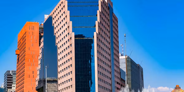 Tall modern office buildings with reflective glass windows and geometric shapes stand against a clear blue sky, with a water fountain visible in the foreground.