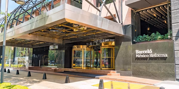 The entrance of Barceló México Reforma hotel features gold-framed revolving doors, a glass and metal canopy, and the hotel name displayed on a black wall to the right.