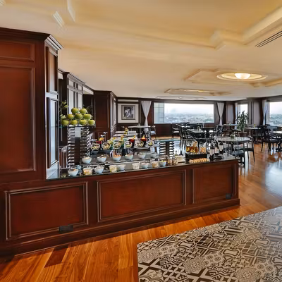 A hotel dining area with wooden floors and paneling, a buffet counter with various breakfast foods, glass jars, and green apples, and tables and chairs arranged near large windows with a city view.