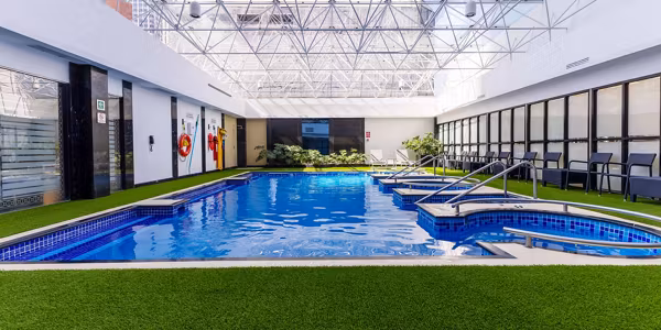 Indoor swimming pool with blue water, surrounded by artificial green grass, lounge chairs, and large windows. Overhead, a glass roof lets in natural light, creating a bright and inviting atmosphere.