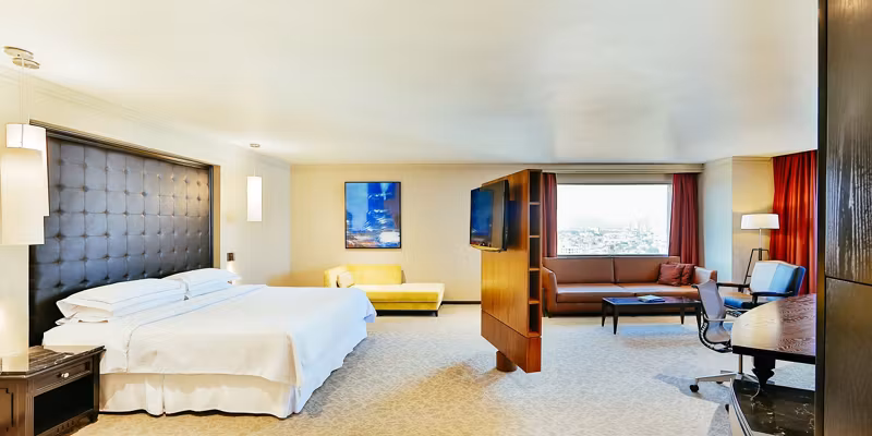 Spacious hotel room with a large bed, padded headboard, modern furnishings, TV divider, sofa, armchair, desk, and a large window letting in natural light.