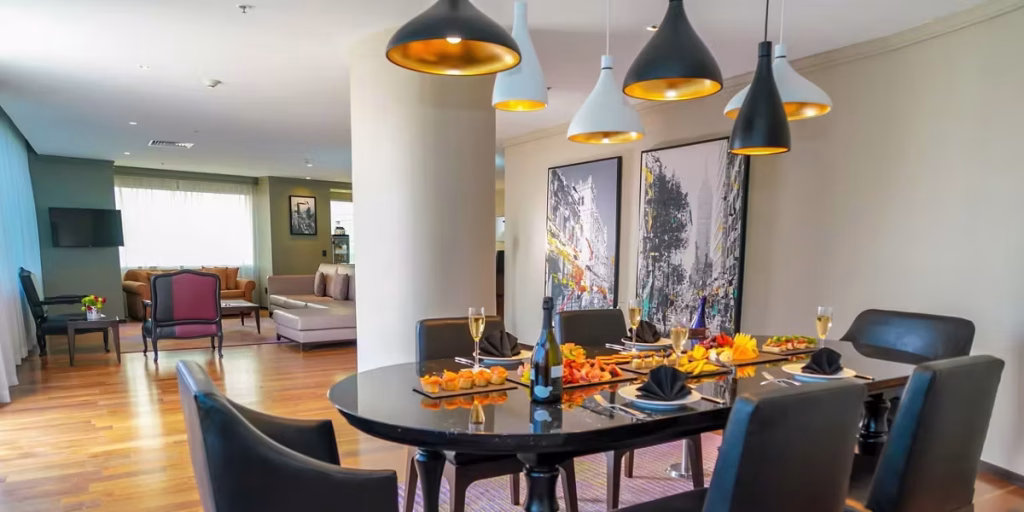 A modern dining room with a black table set for six, featuring plates of appetizers and champagne. Stylish pendant lights hang above, and abstract art is displayed on the wall. A living area with sofas is visible in the background.