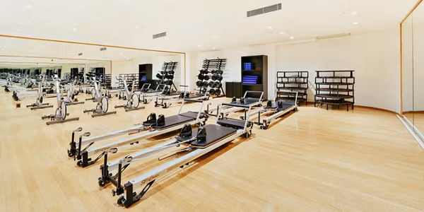 A spacious, brightly lit gym with wooden floors, several pilates reformer machines, weight racks, exercise balls, and large mirrors along the walls. The gym appears clean and organized.
