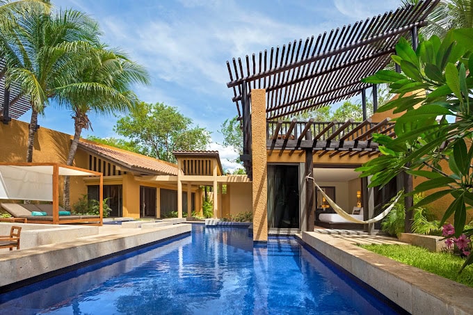 A modern tropical villa features a private blue-tiled pool, a hammock, sun loungers, lush greenery, and shaded pergolas under a bright blue sky.