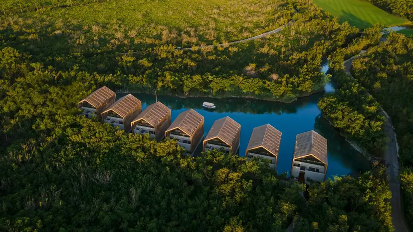 Aerial view of modern villas with slanted roofs built over a narrow blue waterway, surrounded by dense green forest and vegetation. A small boat floats on the water near the villas.