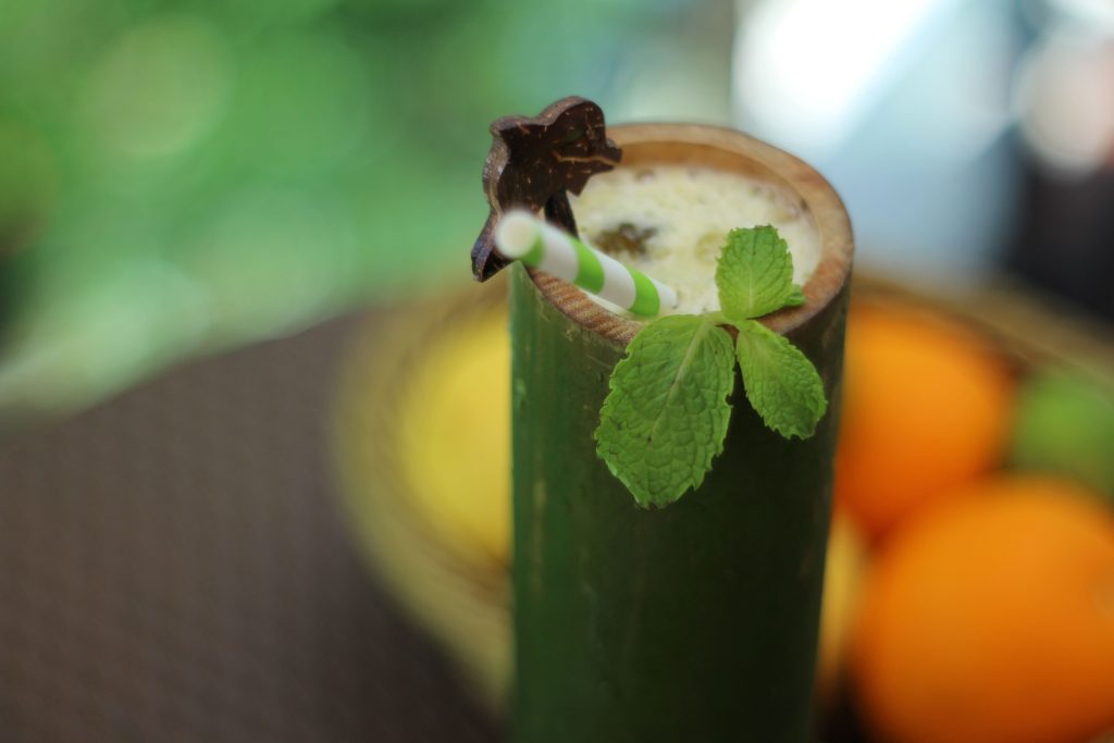 A green bamboo cup filled with a creamy drink, garnished with mint leaves, a green-and-white striped straw, and a star-shaped fruit slice, with blurred oranges and lemons in the background.
