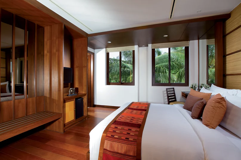 A modern hotel room with a large bed decorated with brown and red pillows and a patterned runner. The room features wooden floors, paneling, a desk, and windows with views of lush green palm trees outside.