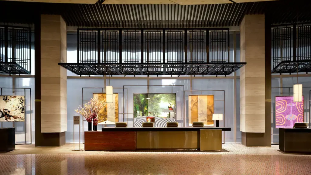 Modern hotel reception area with a sleek, long desk, decorative lamps, and vibrant floral arrangement. Large artworks and a grid-like architectural feature are displayed behind the desk, creating a stylish, inviting atmosphere.