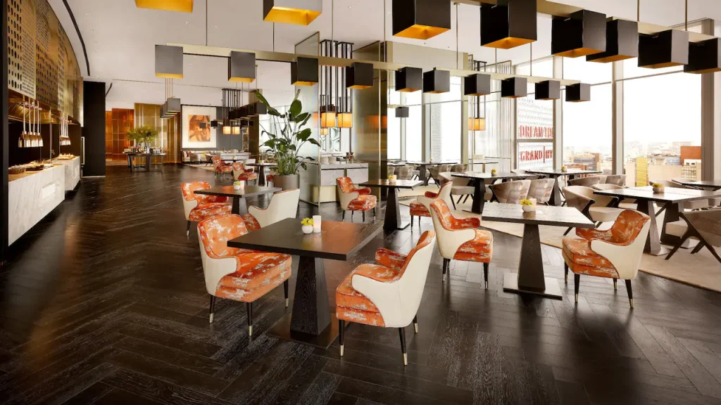 A modern, spacious restaurant with large windows, dark wood floors, patterned orange and white chairs, square tables, and geometric ceiling lights. Buffet stations line the left wall; city views are visible outside.