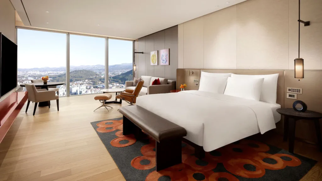 A spacious modern hotel room with a large bed, a dark bench at the foot, a lounge chair, a small table with flowers, and floor-to-ceiling windows offering a panoramic city and mountain view.