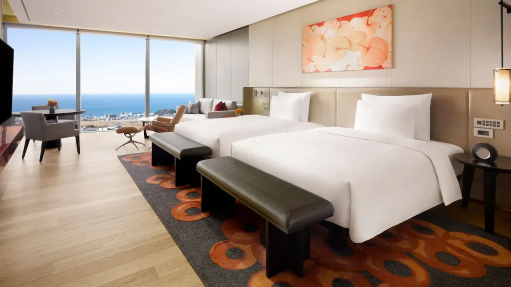 A modern hotel room with two double beds, a patterned carpet, benches at the foot of the beds, large floor-to-ceiling windows offering an ocean view, a work desk, and abstract artwork above the beds.