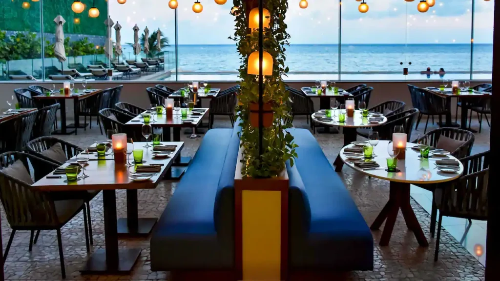 A modern seaside restaurant with tables set for dining, blue cushioned seating, hanging lights, and large windows offering a view of the ocean and palm trees at sunset.