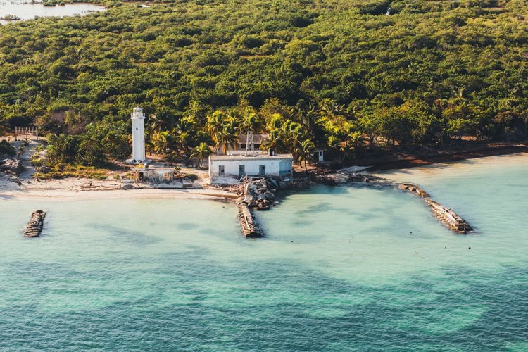Aerial view of a small, white lighthouse and building on a sandy beach with clear turquoise water, surrounded by dense green vegetation and old wooden piers extending into the sea.