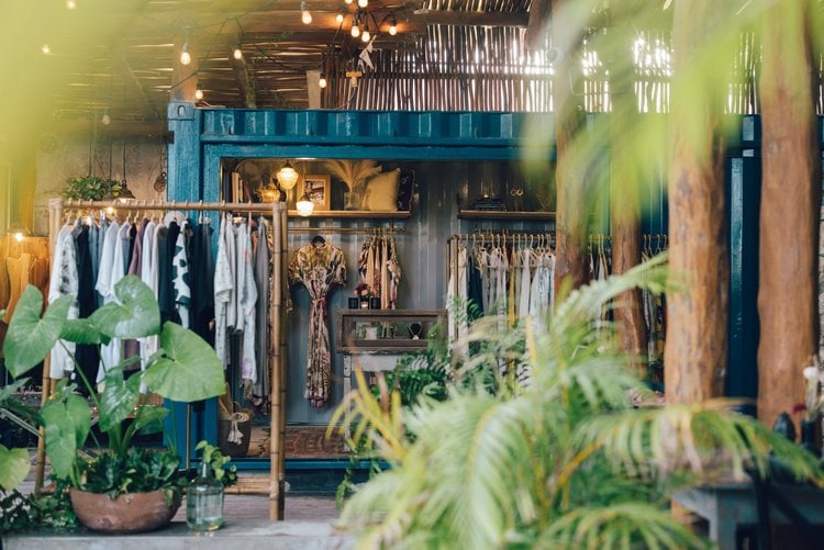 A cozy boutique shop with clothes hanging on racks, surrounded by green potted plants and warm string lights, creating a relaxed, inviting atmosphere.