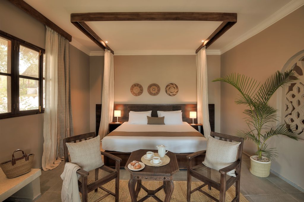 A cozy, neutral-toned bedroom with a four-poster bed, wooden chairs around a table set with tea and snacks, a potted plant, woven wall decor, and natural light from a window with sheer curtains.