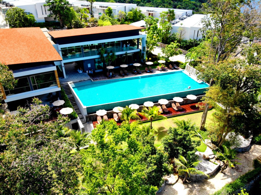 Aerial view of a modern resort featuring a large rectangular swimming pool with lounge chairs and umbrellas, surrounded by lush green trees and landscaped gardens. White buildings are visible in the background.