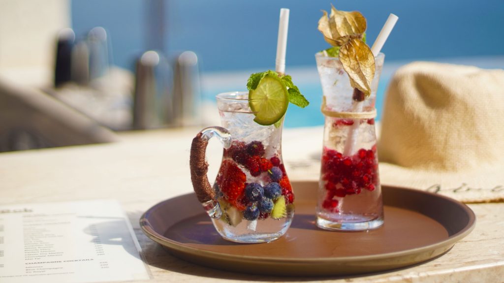 Two refreshing drinks with ice, berries, and lime slices are served in clear glasses with straws on a brown tray. A menu and a sun hat are on the table, with a blurred outdoor background.
