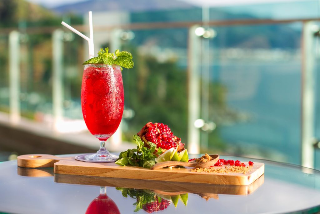 A red iced drink with mint and two straws sits on a glass table next to a wooden board with pomegranate, lime, and garnishes. A blurred outdoor background with glass railings and hills is visible.