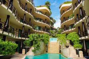 Curved, multi-story balconies with black railings overlook a lush courtyard featuring a swimming pool and a stone waterfall, surrounded by tropical plants and statues, under a blue sky.