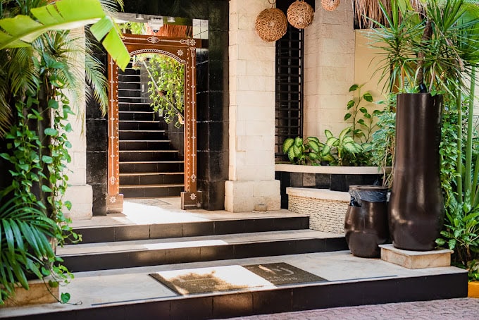 A tropical hotel entrance with lush green plants, black and white stairs, a decorative wooden archway, and woven hanging lamps. Sunlight brightens the scene, creating a welcoming atmosphere.