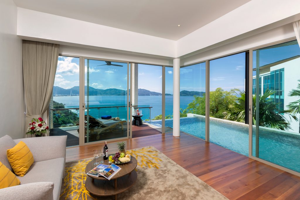 Modern living room with a gray sofa, yellow pillows, and a rug, opening to a terrace with a private pool and panoramic ocean and mountain views through large glass doors. A tray with wine and fruit is on the coffee table.