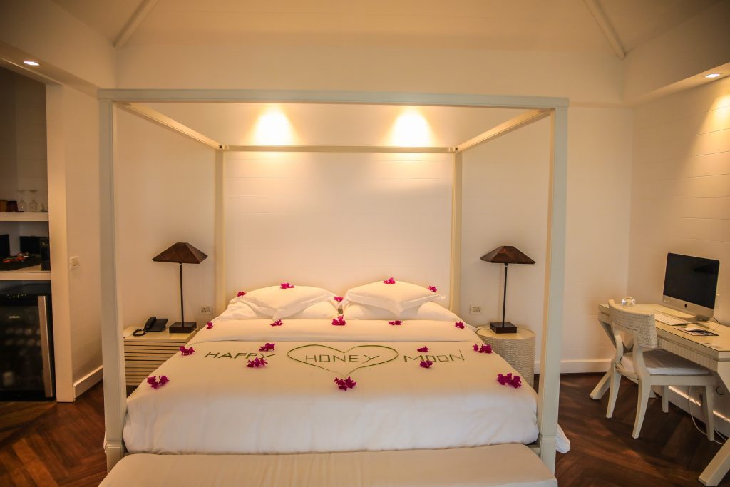 A cozy hotel room with a canopy bed decorated with flower petals spelling Happy Honeymoon. Two bedside tables with lamps are on either side, and a desk with a computer sits in the corner.