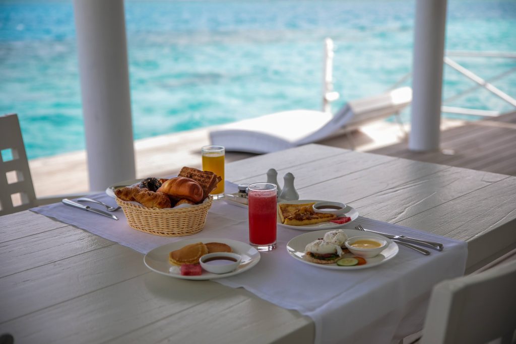 A white table set for breakfast overlooks turquoise water. Plates of eggs, pastries, fruit, and juice glasses are arranged neatly, with a basket of bread in the center and sunlight streaming onto the deck.